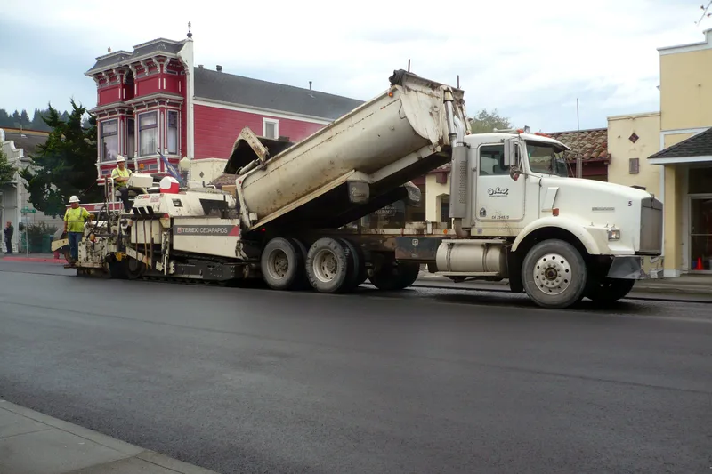 Dump truck menuangkan aspal hotmix panas ke dalam hopper mesin asphalt paver finisher beroda rantai untuk penghamparan jalan heavy duty