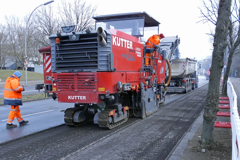 Unit mesin cold milling Wirtgen beralas roda rantai baja sedang mengupas lapisan aus aspal rusak dan menyalurkan material gilingan RAP ke bak dump truck pada proyek pemeliharaan jalan raya