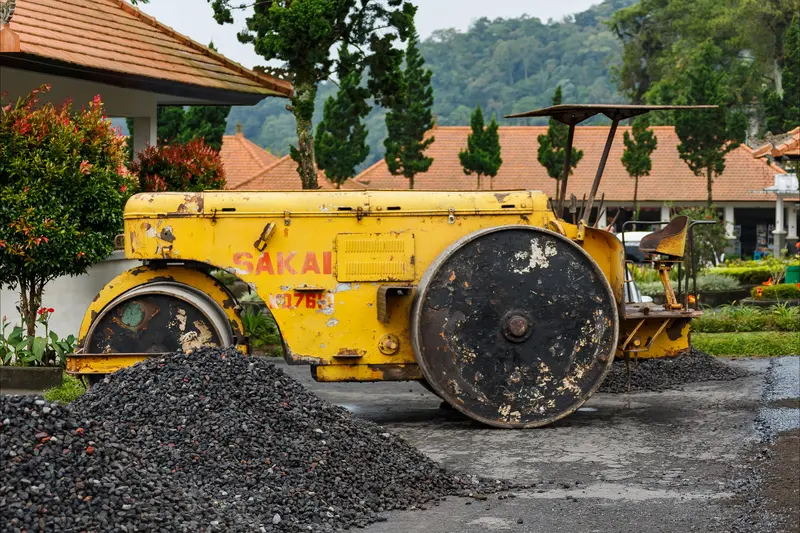 Mesin pemadat tandem roller Sakai dan tumpukan agregat batu pecah di sisi badan jalan pegunungan beraspal dengan latar perbukitan tropis berkabut