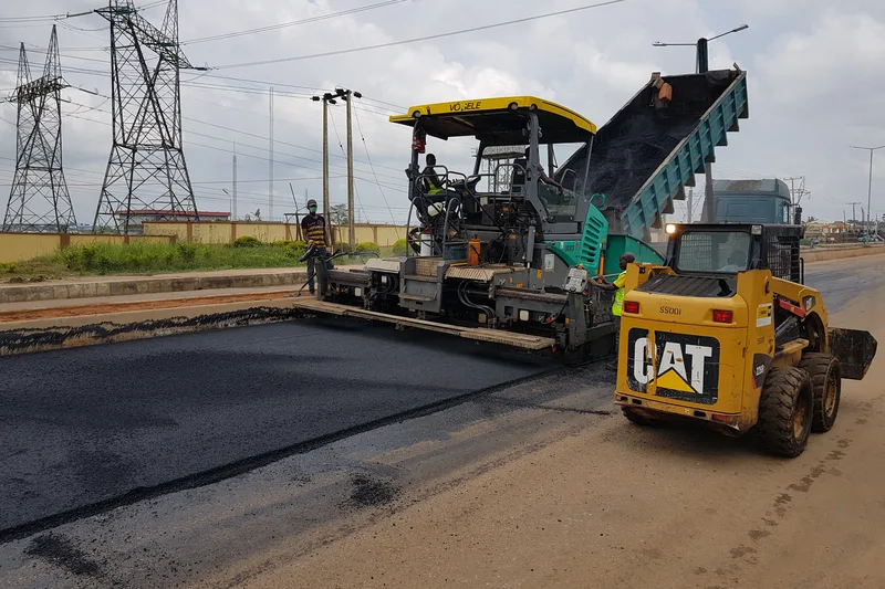 Proses penghamparan aspal hotmix menggunakan mesin asphalt paver Vögele dan dump truck pengangkut material panas pada proyek peningkatan jalan perbukitan