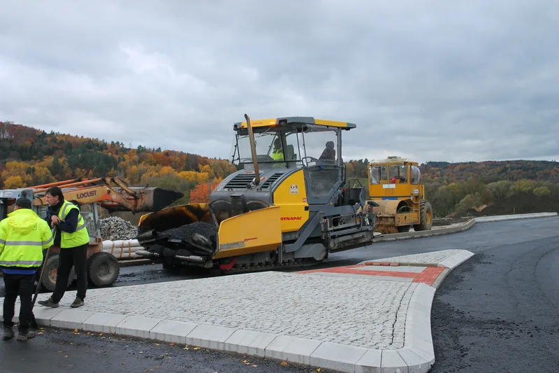 Unit mesin asphalt paver crawler dan tandem roller compactor sedang menggelar serta memadatkan aspal hotmix pada persimpangan jalan berkerb beton